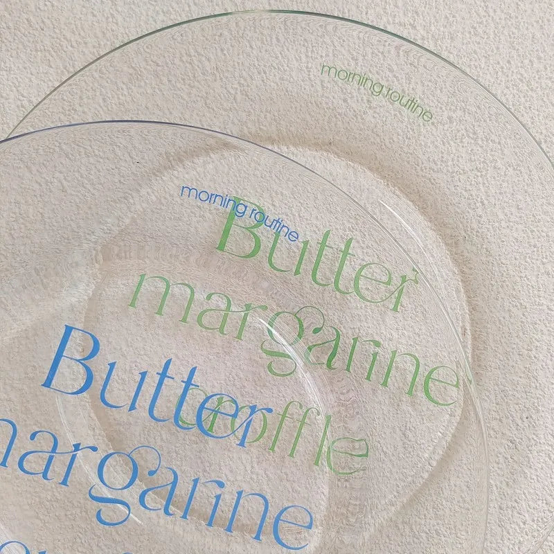 Two clear glass plates with printed text on a textured cream background. The front plate is printed with 'Butter margarine' in blue and green, and the back plate reads 'morning routine' in green.