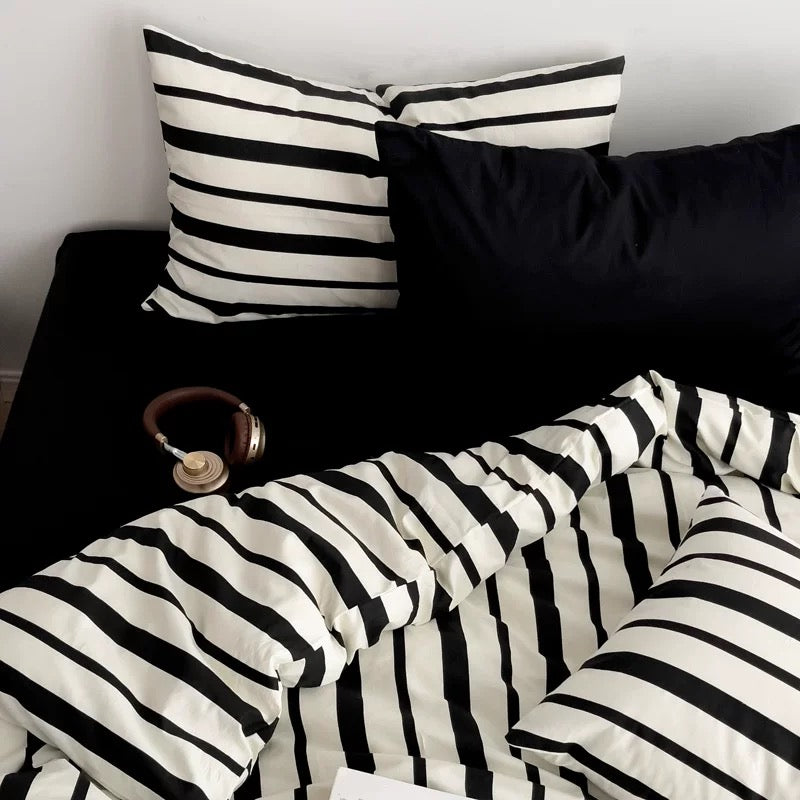 Black and white striped bedding set with matching pillows and duvet, styled on a bed with a black headboard, a black pillow, and a pair of brown over-ear headphones.