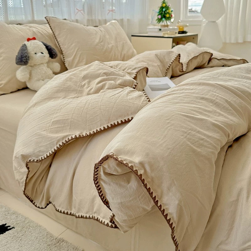 Beige comforter set on a bed with decorative pillows and a small tree in the background.