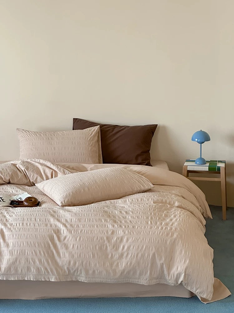 A neatly made bed with textured cream and beige bedding, two dark brown pillows, and a small wooden bedside table with a blue lamp in a minimalist bedroom setting.