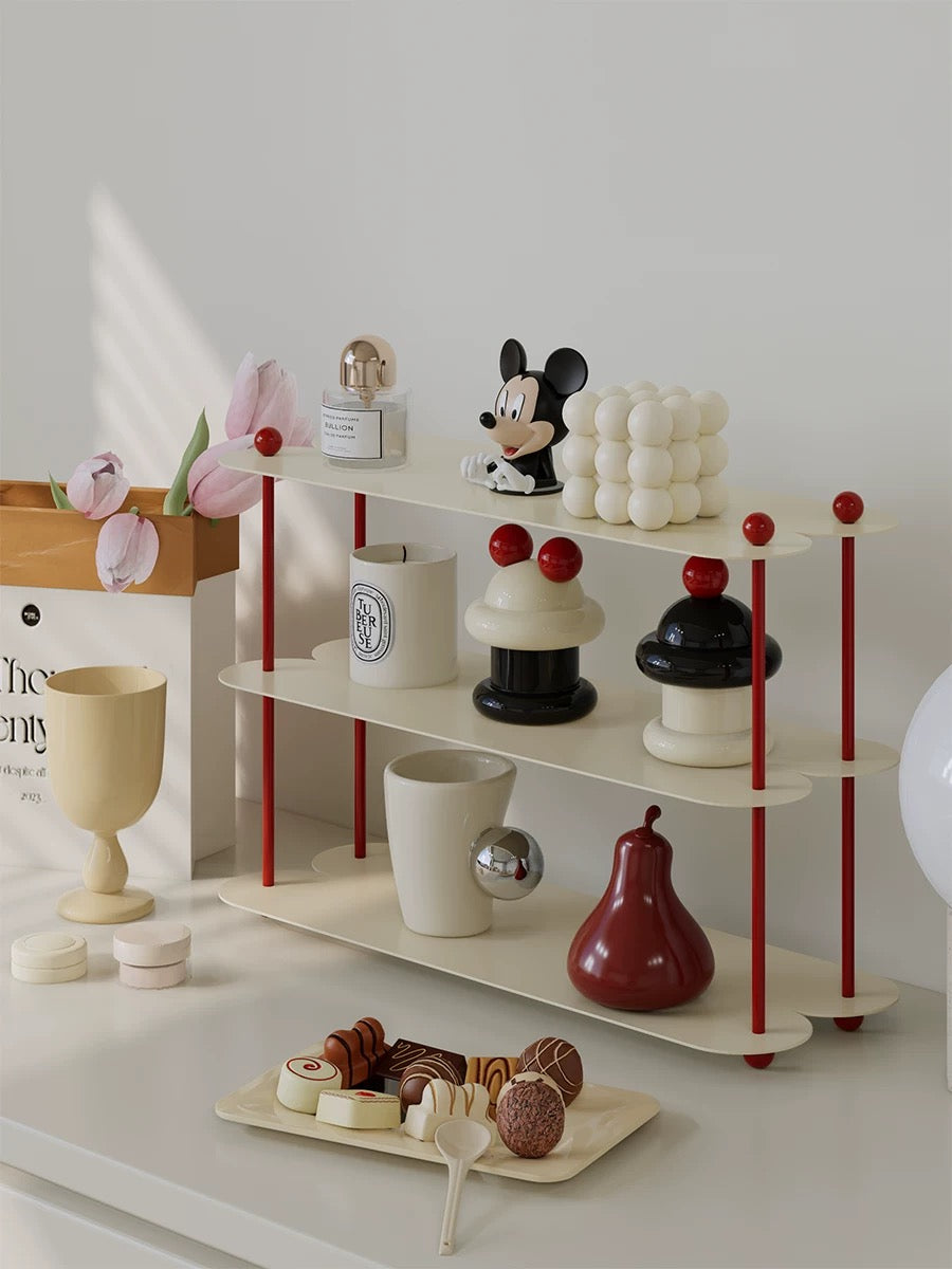 A cream coffee mug shelf with red posts displays candles, a mug and decorative objects on three levels.