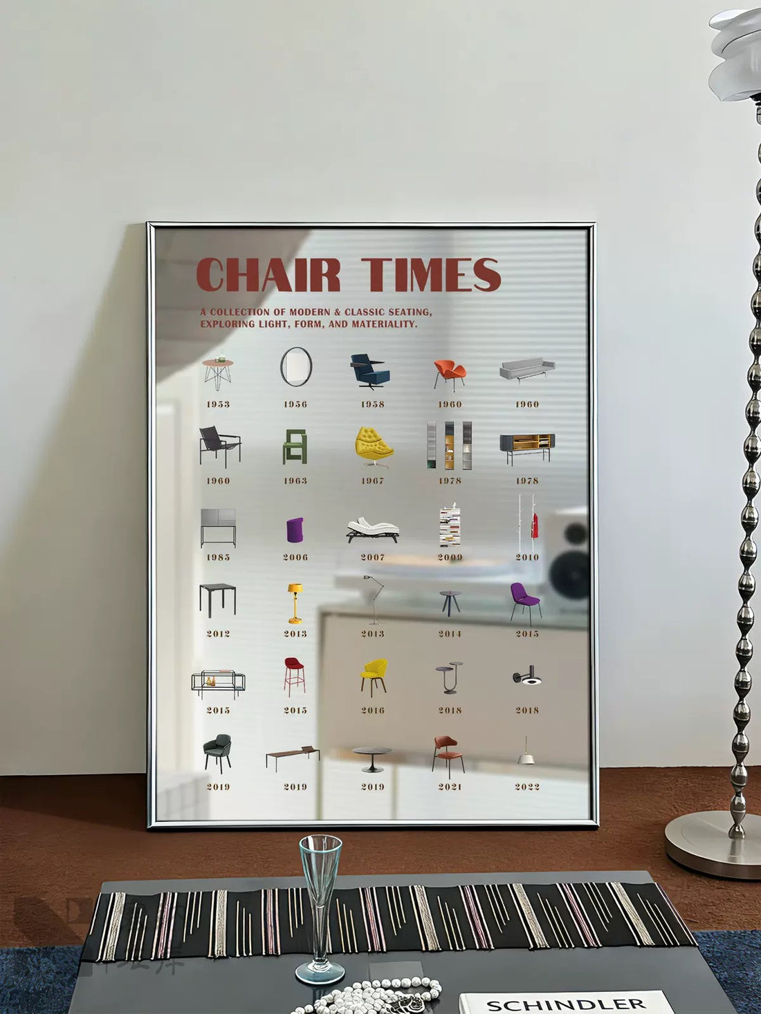 A framed art print featuring a collage of small furniture icons and letters, displayed on a white shelf in a styled room next to a person.