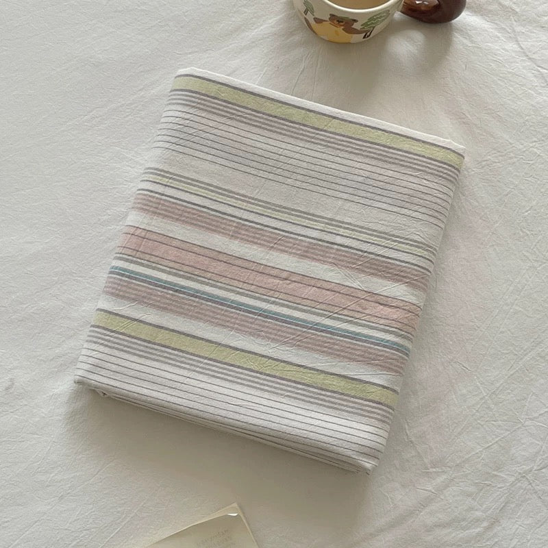 A folded cotton muslin blanket with pastel horizontal stripes in pink, blue, yellow, and gray on a white background, styled on a white fabric surface with a small ceramic cup in the background.
