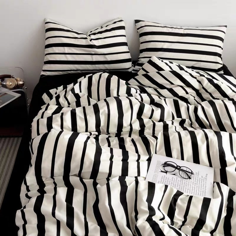 A cozy bed setup featuring a black and white striped bedding set, including a duvet cover and matching pillowcases, styled with a black pillow, a bedside table, and a magazine.