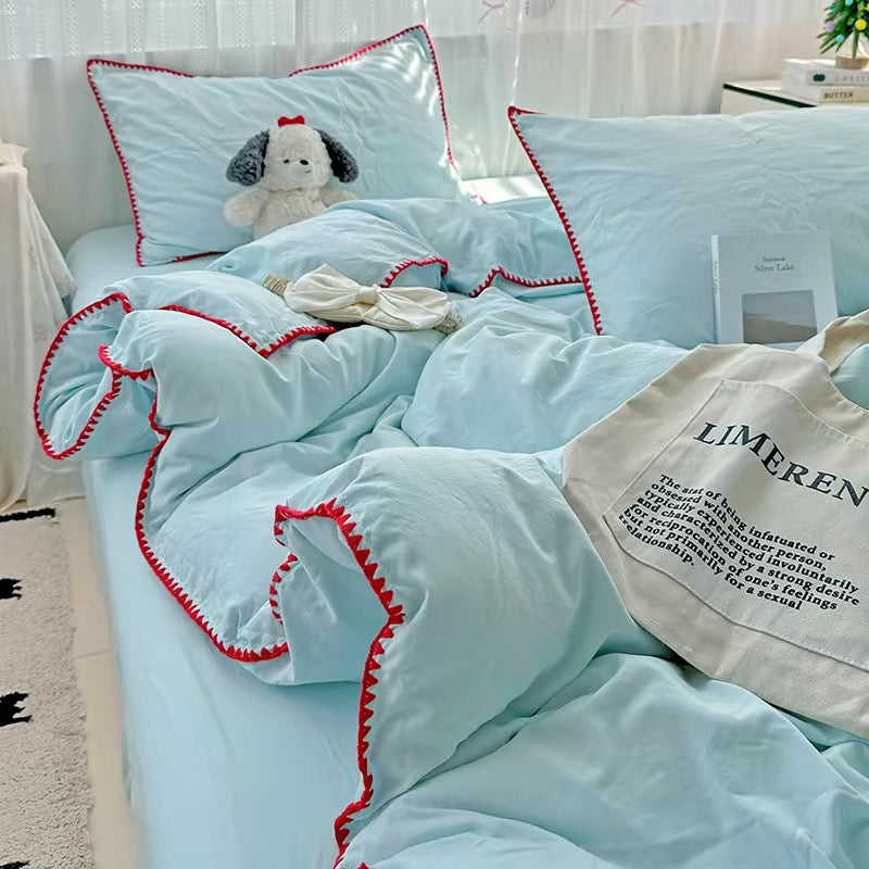 Light blue bedding set with red trim on a bed, featuring a teddy bear and a bag.