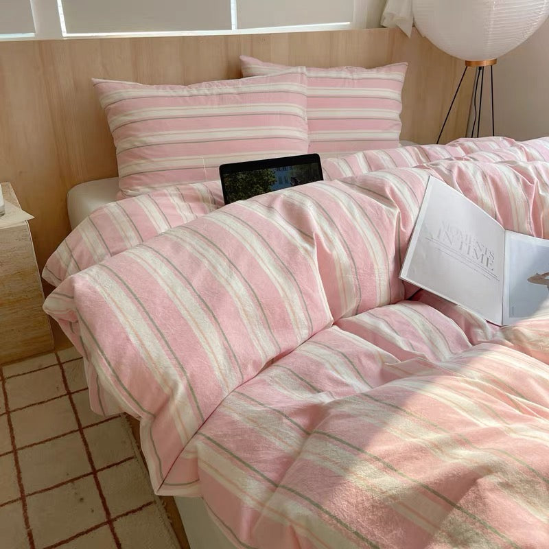 Pink and white striped bedding set on a bed with a nightstand and lamp in the background.