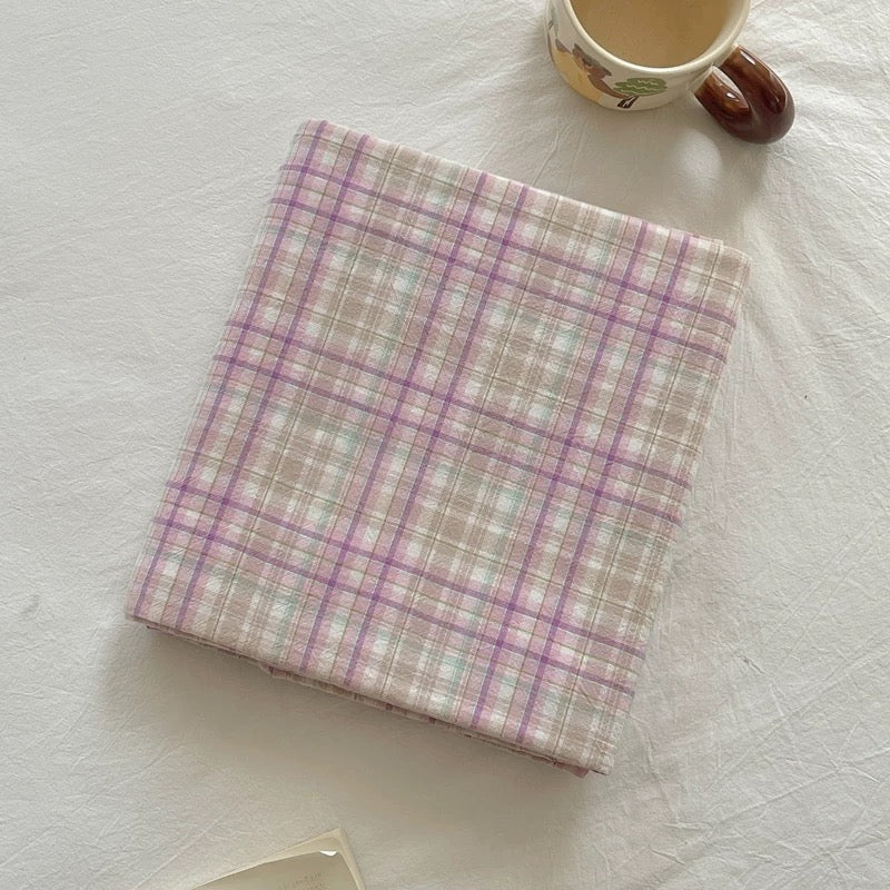 A folded square fabric item, likely a napkin or small cloth, featuring a pastel plaid pattern in pink, purple, and beige, styled on a white textured fabric surface next to a ceramic mug.