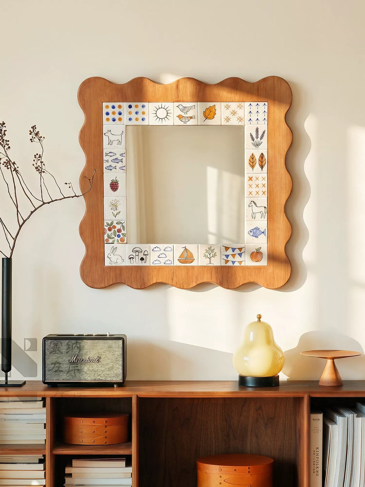 A Cottagecore mirror with a wavy brown frame hangs above a cabinet beside a radio and a yellow lamp.