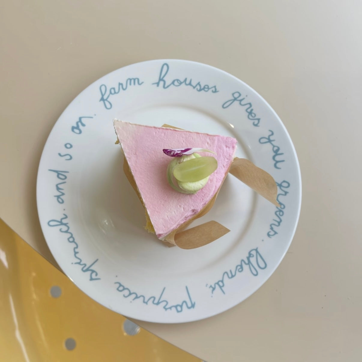 A triangular slice of pink cake with a green pear garnish sits on a white ceramic plate with blue cursive text, set against a beige background with a yellow shape in the lower-left corner.