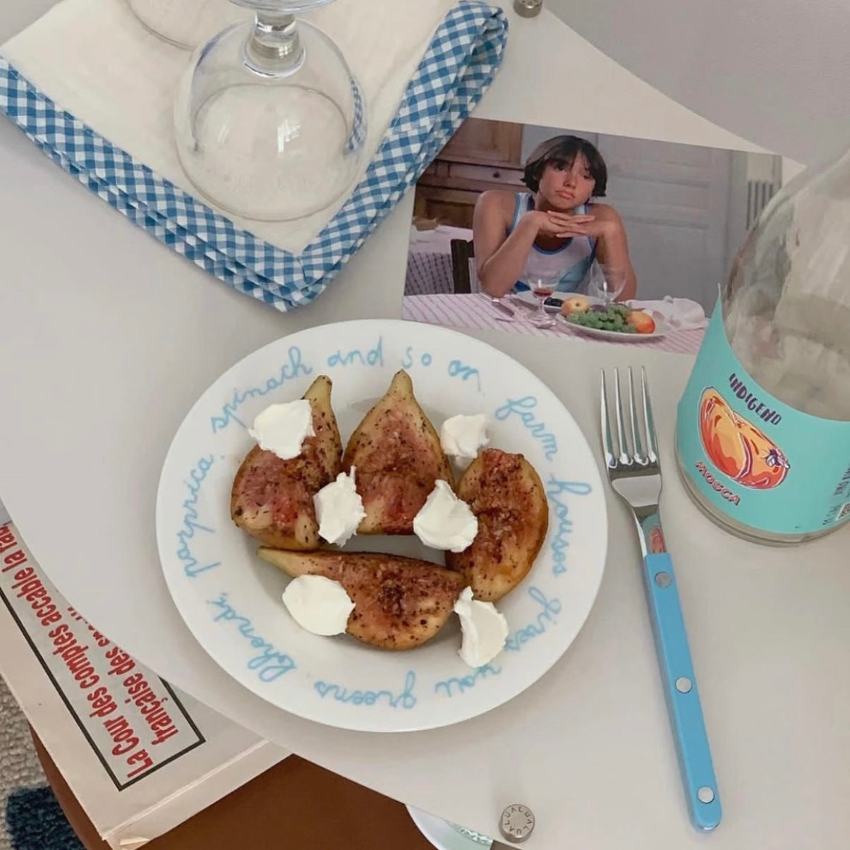 A styled tabletop scene with a white plate holding several baked pears topped with white cream, alongside a blue-handled fork, a glass bottle, a napkin, and a small printed photo of a woman at a table.