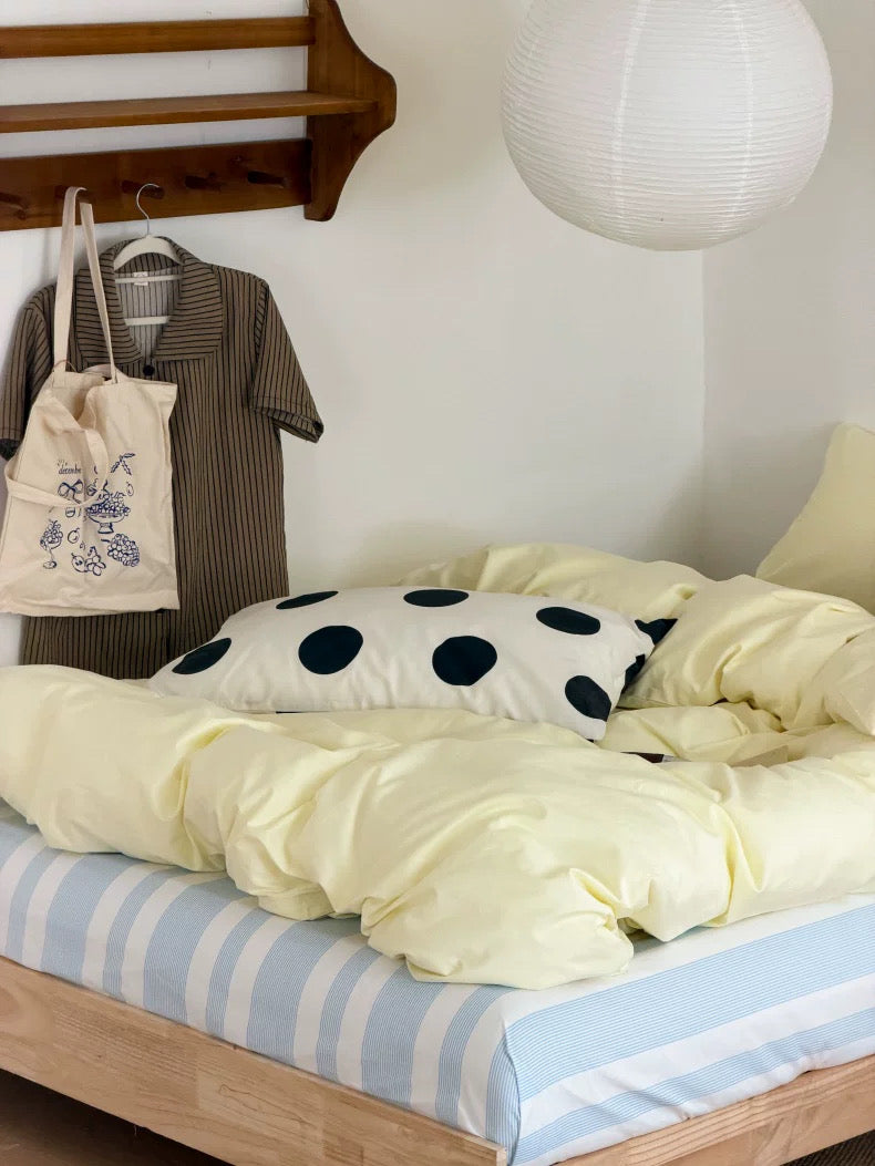 A cozy bedroom scene with a bed made up in pale yellow bedding, a light blue and white striped sheet, a cream pillow with dark navy polka dots, and a wooden wall shelf holding a striped shirt and a canvas tote bag.