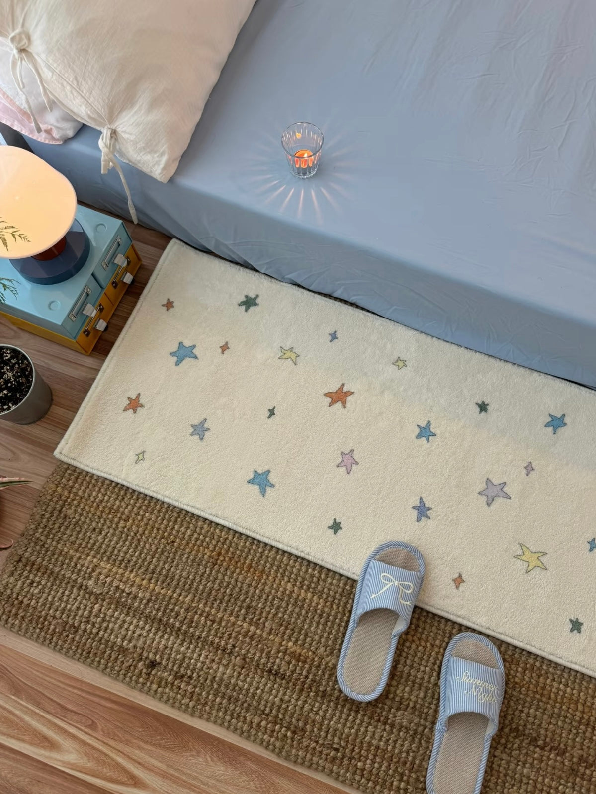 A cream rug with multicolor stars lies flat beside a blue bed, shown from above with slippers nearby.