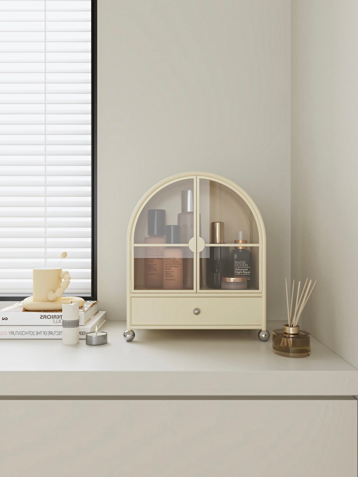 A cream perfume cabinet faces forward, with bottles behind the glass doors and a silver knob below.