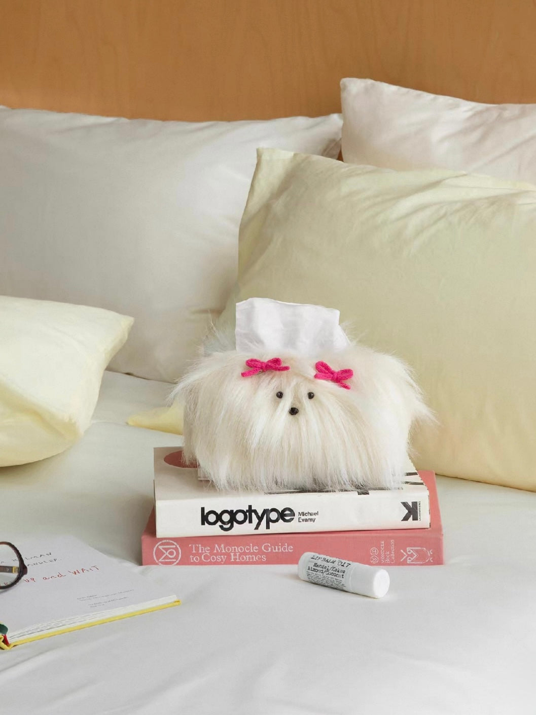 Cream White tissue cover with pink bows rests on stacked books beside a pale green pillow on white bedding.