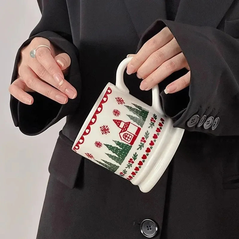 A person in a black blazer holds a white ceramic mug decorated with a red and green Christmas pattern featuring snowflakes, a house, and pine trees.
