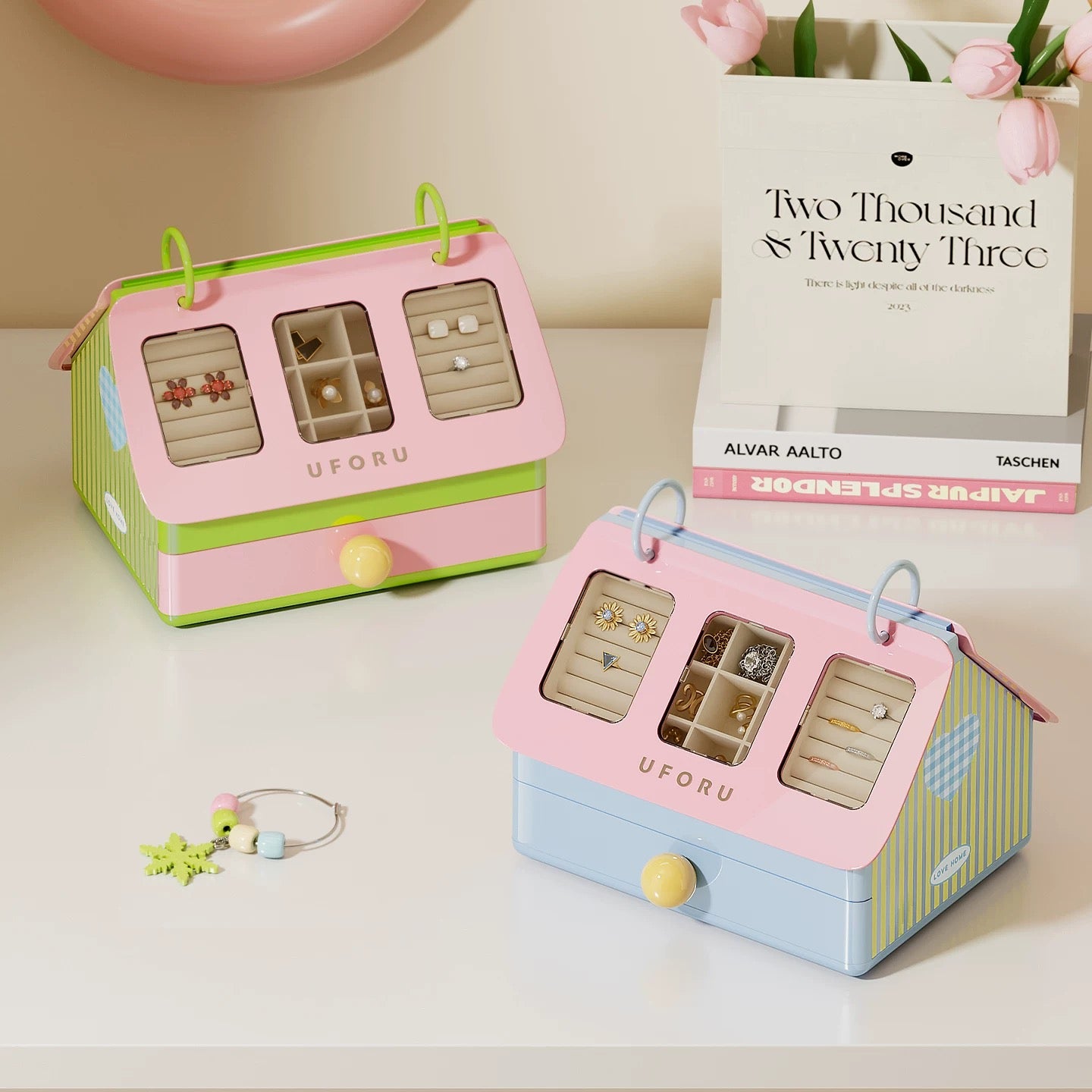 Two small jewelry boxes with compartments on a surface, one pink and green, the other pink and blue, with a book titled 'Two Thousand & Twenty Three' in the background.