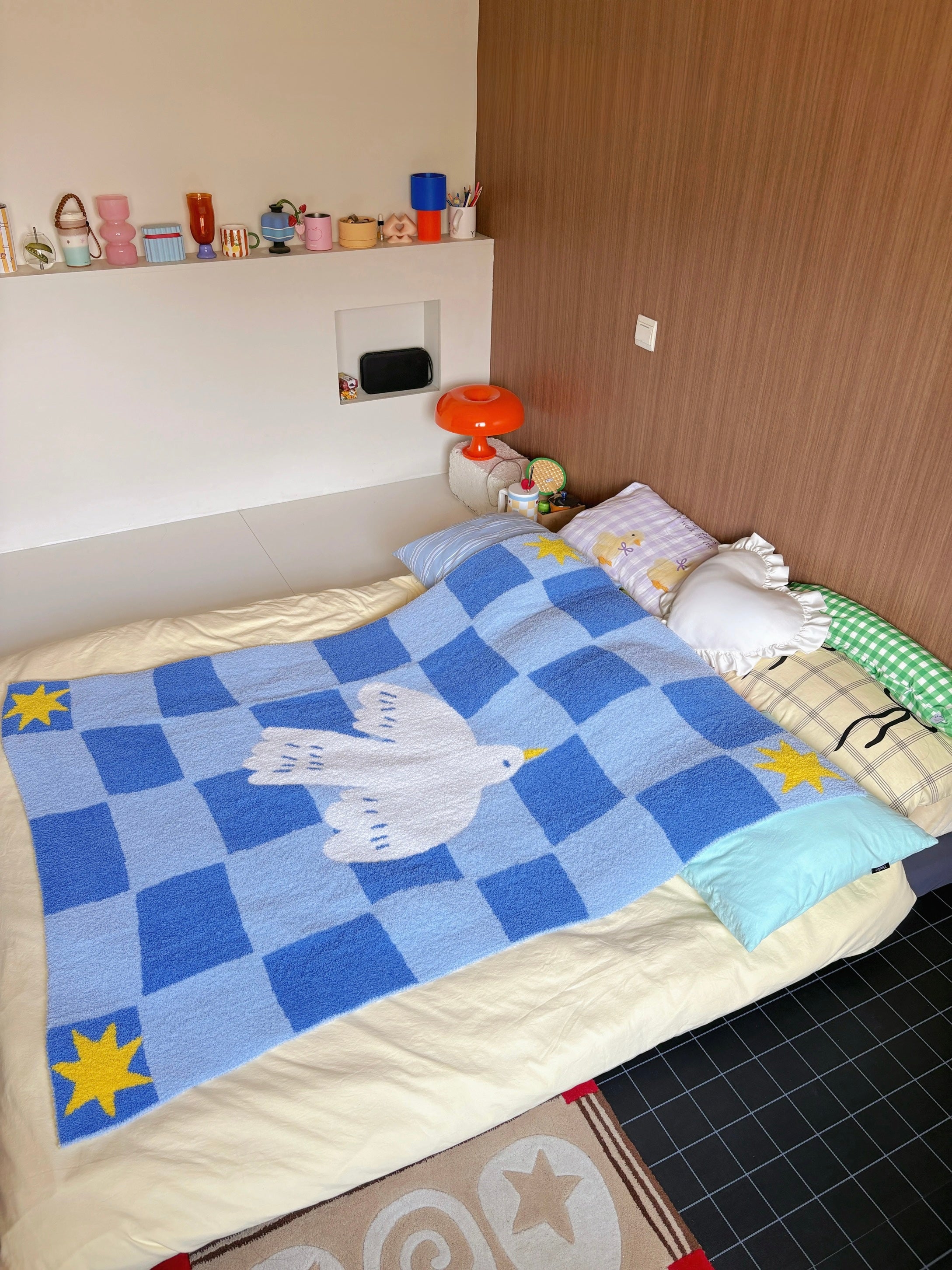 A cozy bedroom scene showing a blue checkered blanket with a white dove in the center and yellow stars in the corners, spread across a bed with cream bedding and decorative pillows.