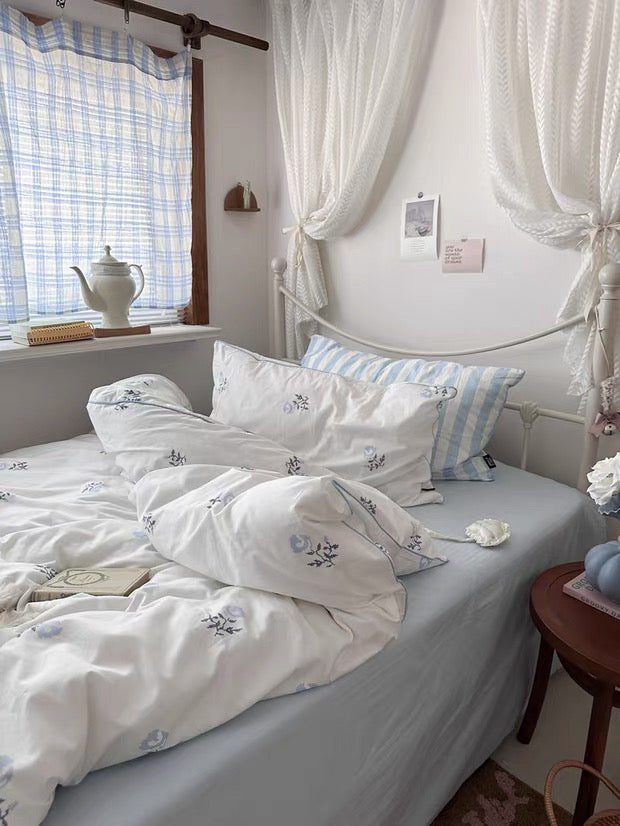 Bedroom with white bedding and floral patterns, curtains, and a teapot on a windowsill.