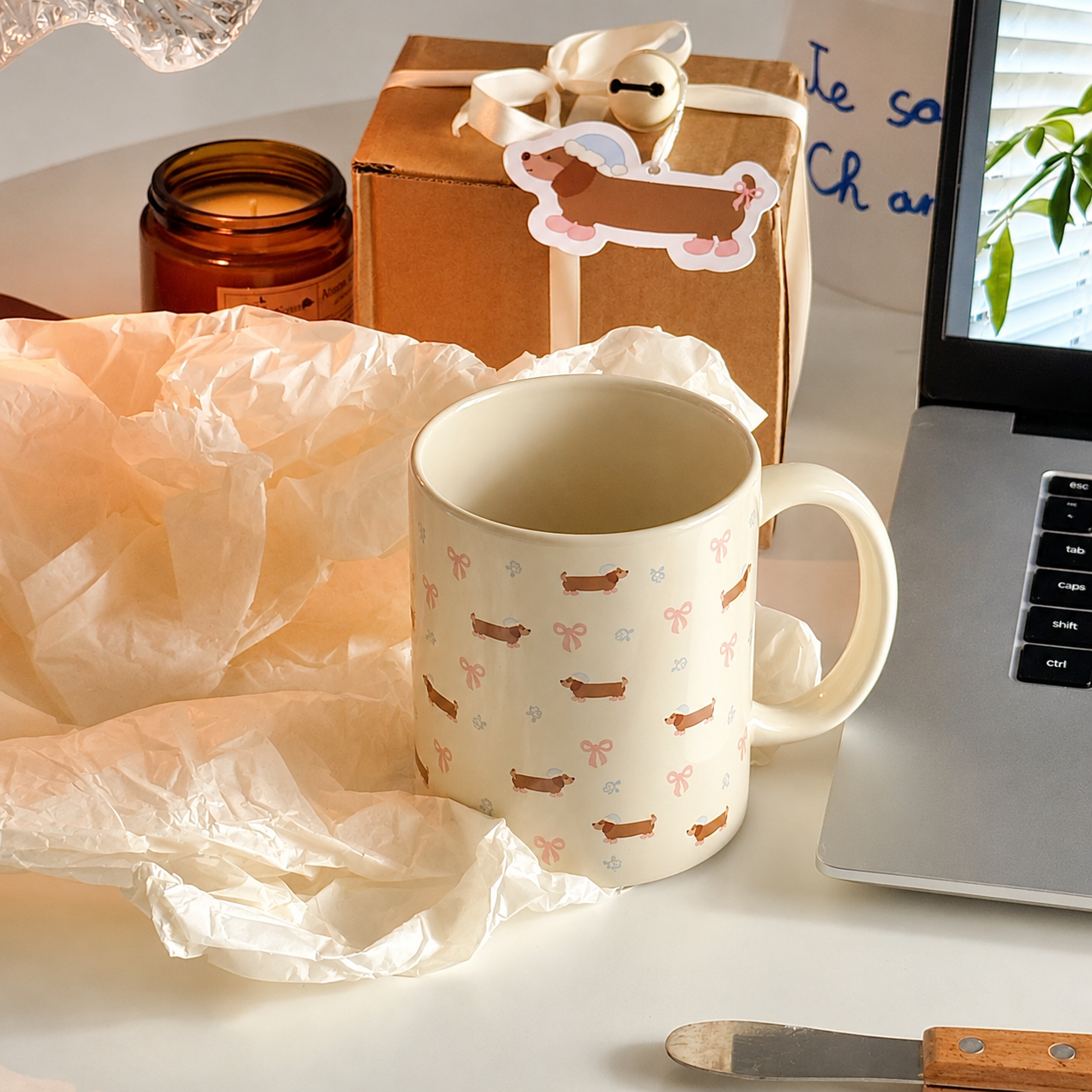 A cream-colored ceramic mug with a pattern of dachshunds and bows sits on a desk next to a laptop, a gift box, and crumpled tissue paper.