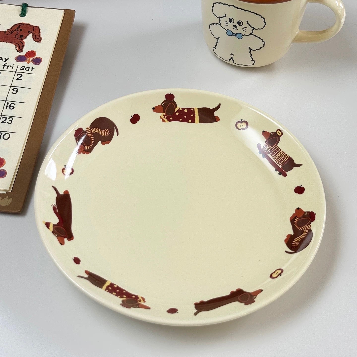 An off-white ceramic plate with a border pattern of dachshund dogs in brown sweaters, shown on a light gray surface next to a mug and a calendar.