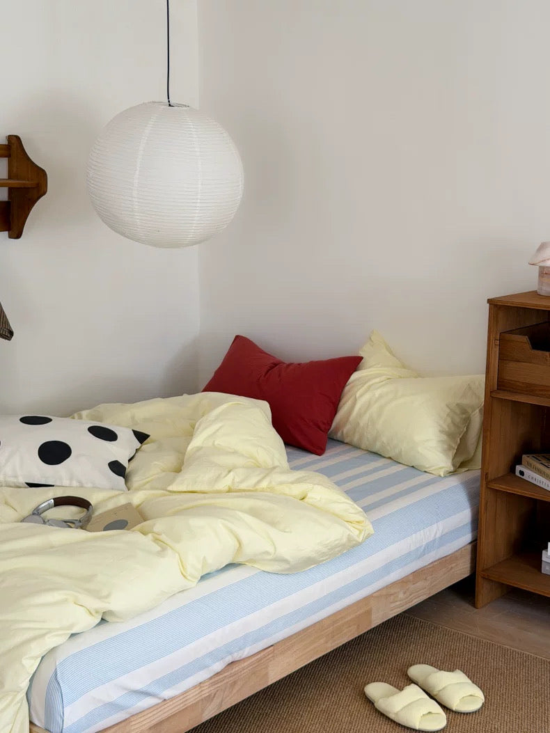 A cozy bedroom scene with a light wood bed frame made up with pale yellow bedding, a blue-and-white striped mattress cover, cream pillows, a red accent pillow, a black-and-white polka dot pillow, a white hanging pendant lamp, and a wooden nightstand.