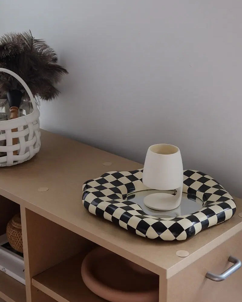 A black and cream checkered woven tray holding a small white table lamp, placed on a light wood cabinet against a gray wall.