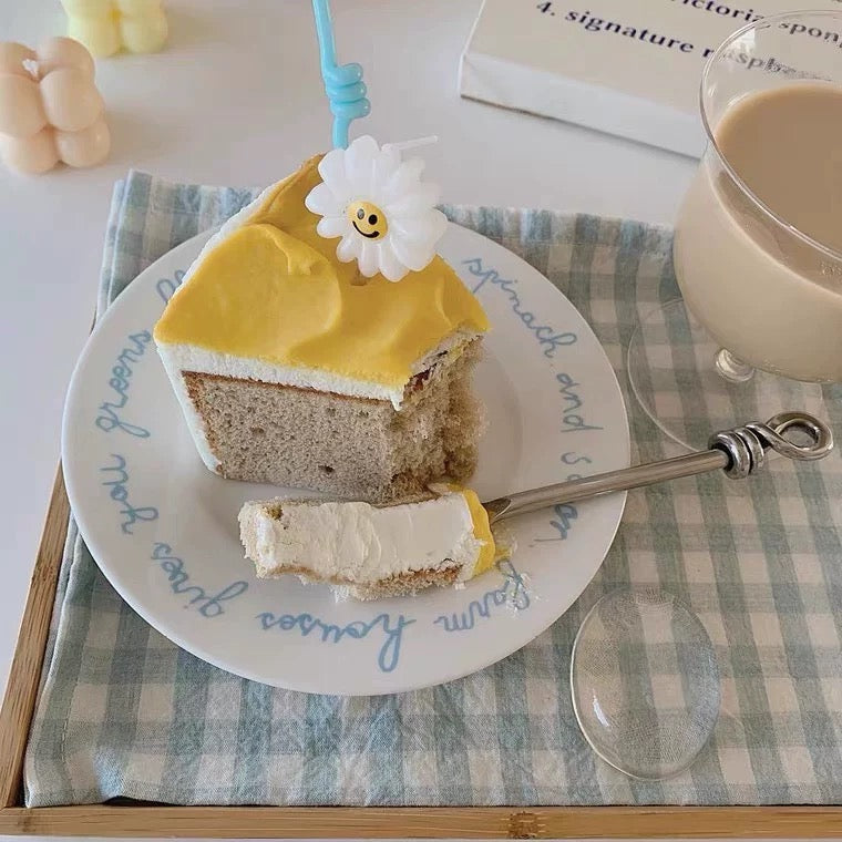 A slice of layered cake on a white plate with blue script text, decorated with a white daisy flower, set on a blue-and-white checkered cloth next to a glass of milk.