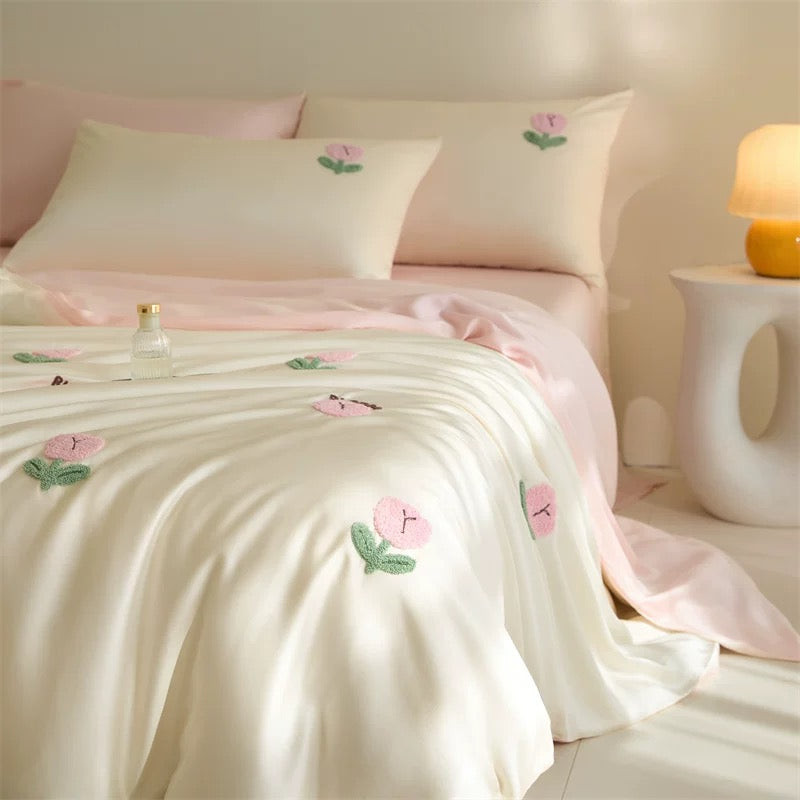 A soft cream-colored satin or silk duvet cover and pillowcases with embroidered pink flowers and green leaves, styled on a bed in a warm, softly lit bedroom.