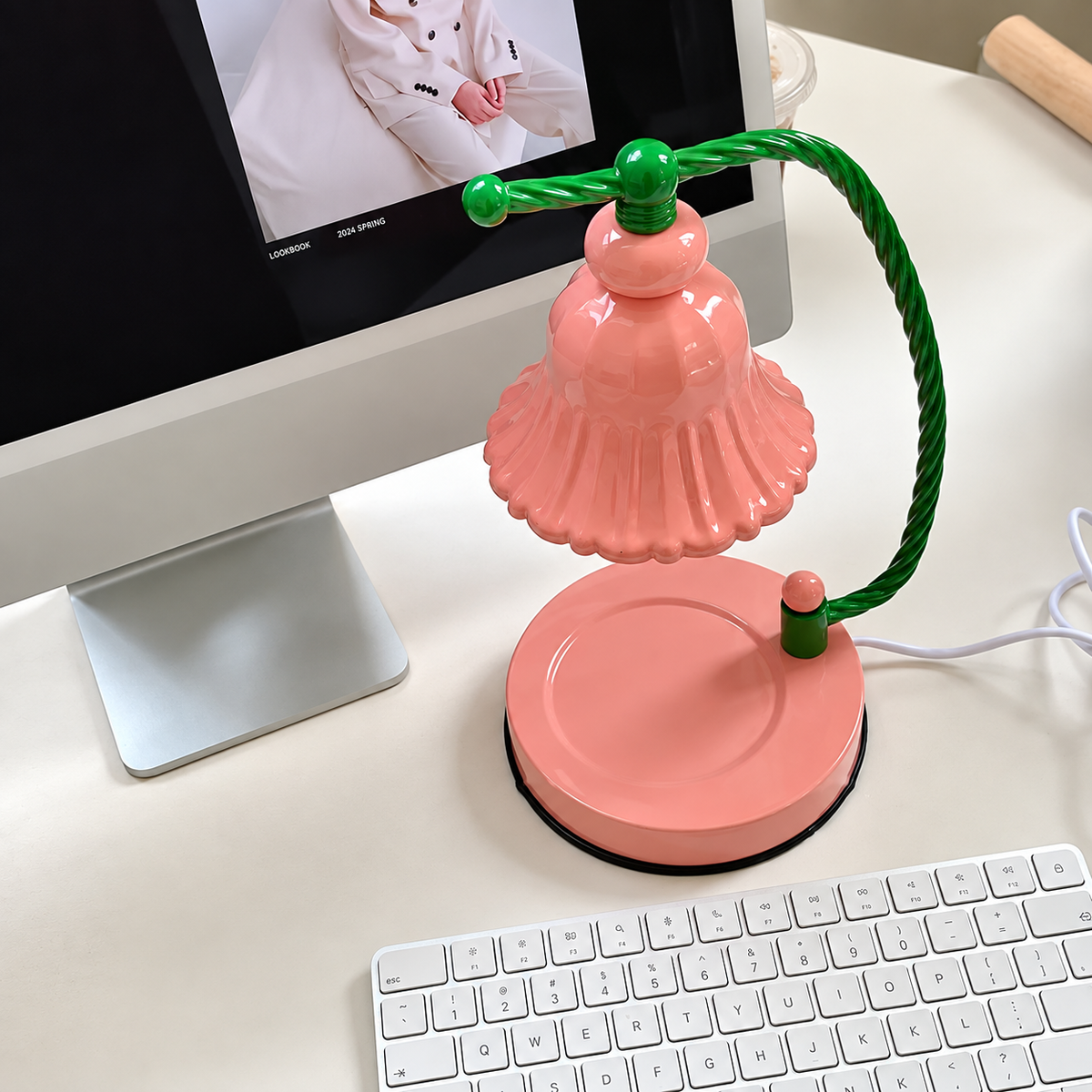 A retro-style desk lamp with a pink bell-shaped shade and base, supported by a twisted green wire arm, shown on a white desk next to a computer monitor, keyboard, and mouse.