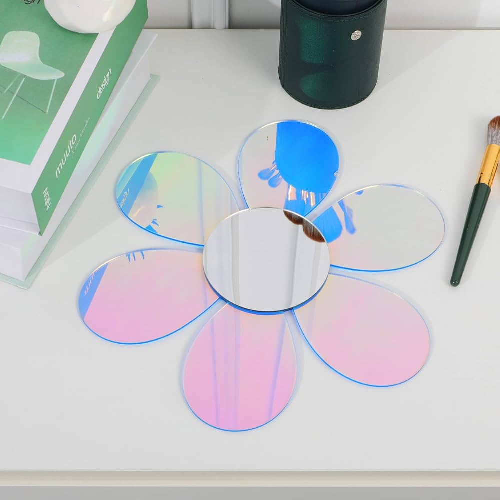 A flower-shaped makeup mirror with iridescent pink, blue, and purple reflective petals and a silver mirror center, displayed on a white tabletop with books, a black container, and a makeup brush nearby.
