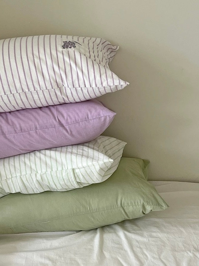 Four pastel pillowcases stacked with lavender grape stripes, dusty lilac, sage flower stripes and solid green.