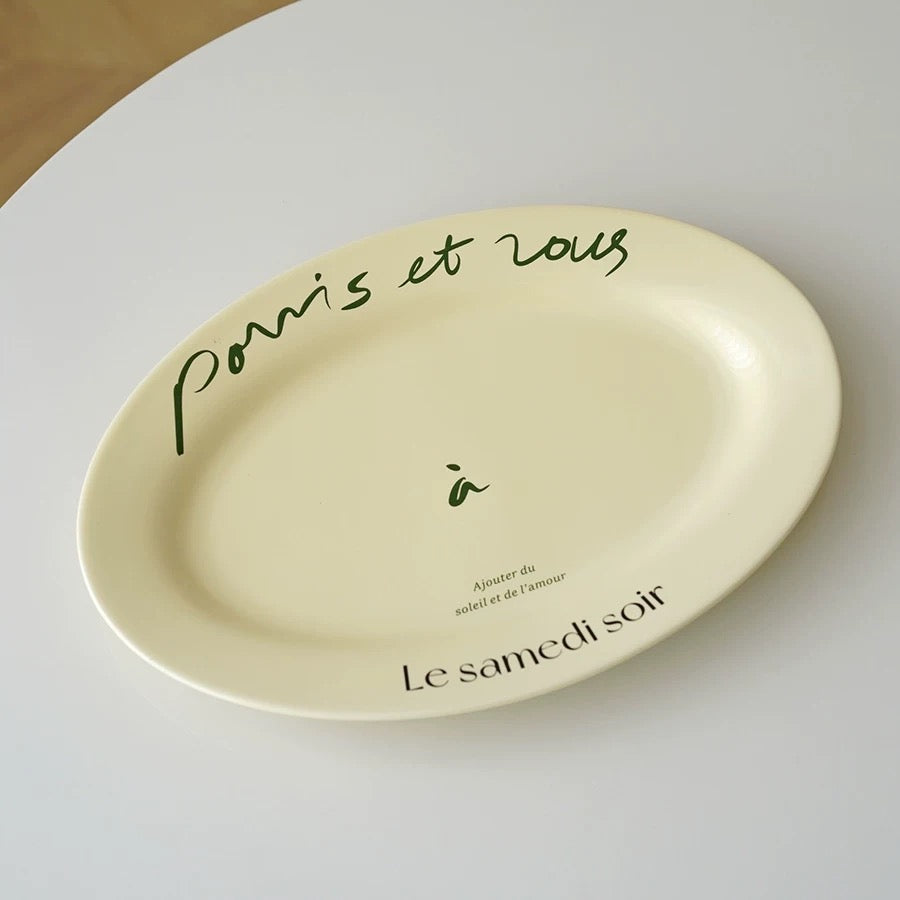 Oval cream porcelain plate with black French lettering photographed from above on a pale background
