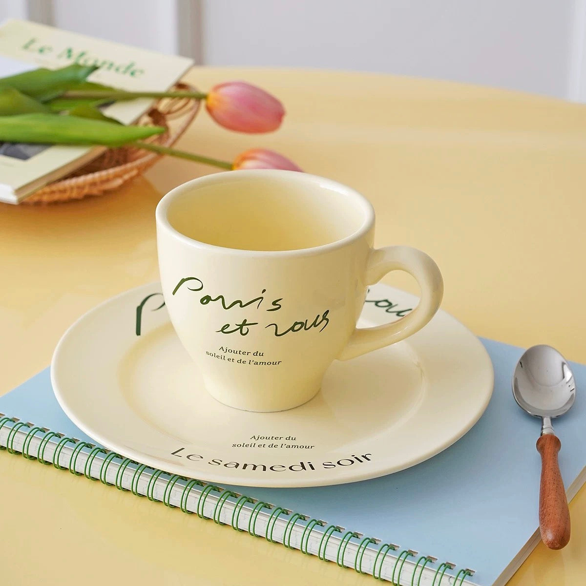 A cream-colored ceramic cup and saucer set with French script text, styled on a light yellow tabletop with a blue notebook, a spoon, and tulips in the background.