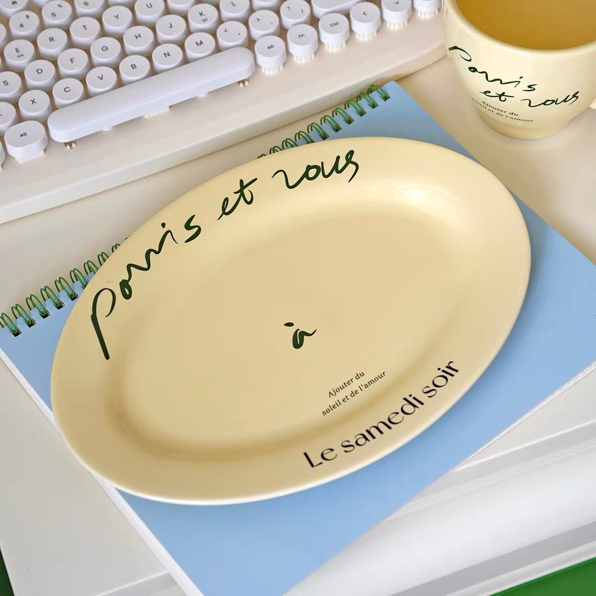 A cream-colored oval ceramic plate with French script text, displayed on a light blue notebook next to a white keyboard and a matching cream mug.