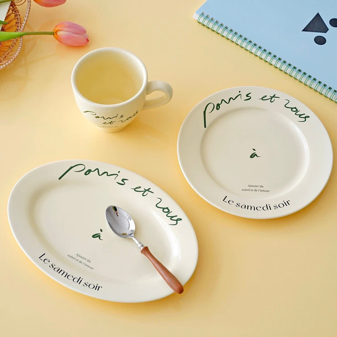 Two round ceramic plates and a matching mug with French script text, styled on a pale yellow surface with a spoon, flowers, and a notebook.