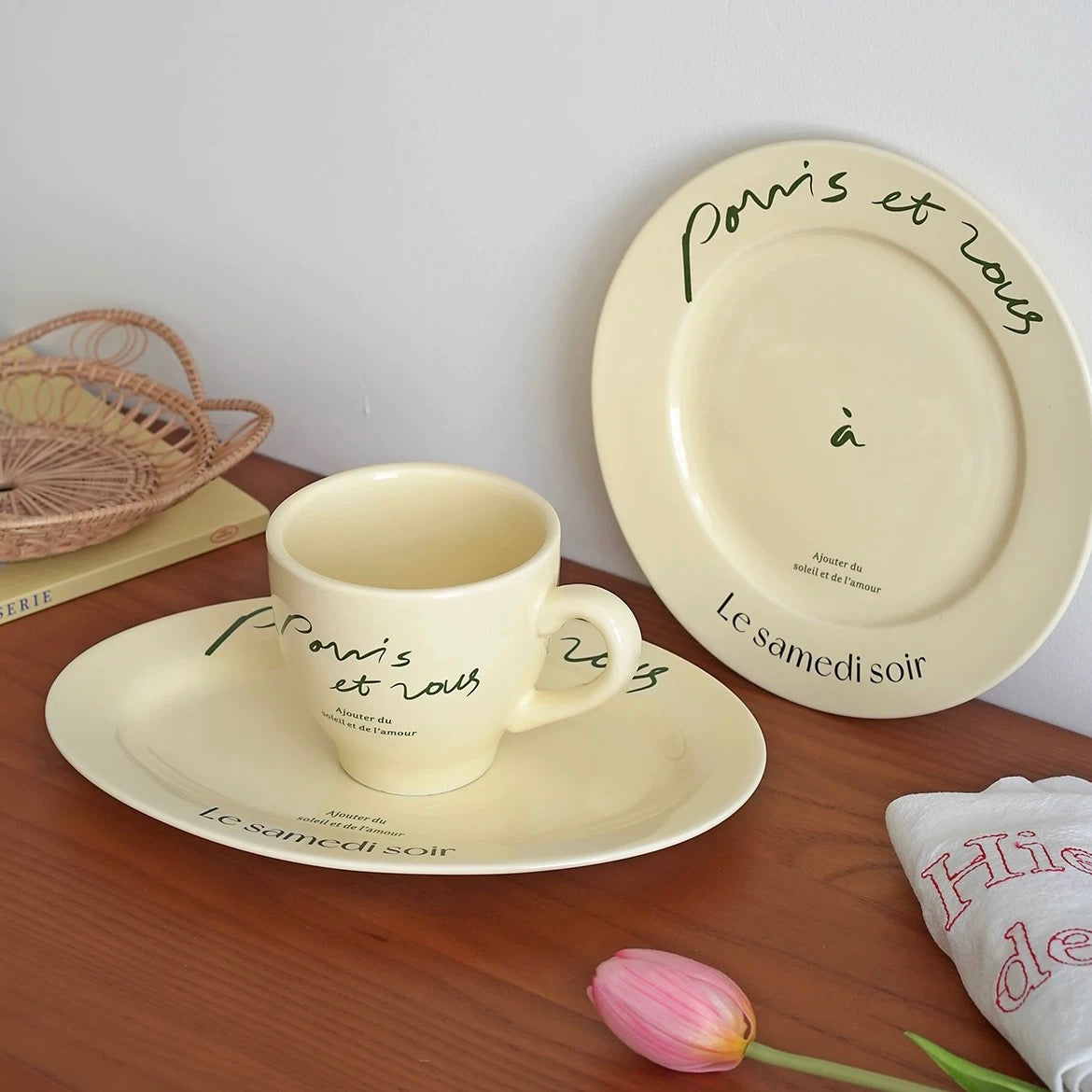 Cream porcelain mug and round plate with black French script displayed together on wooden tabletop
