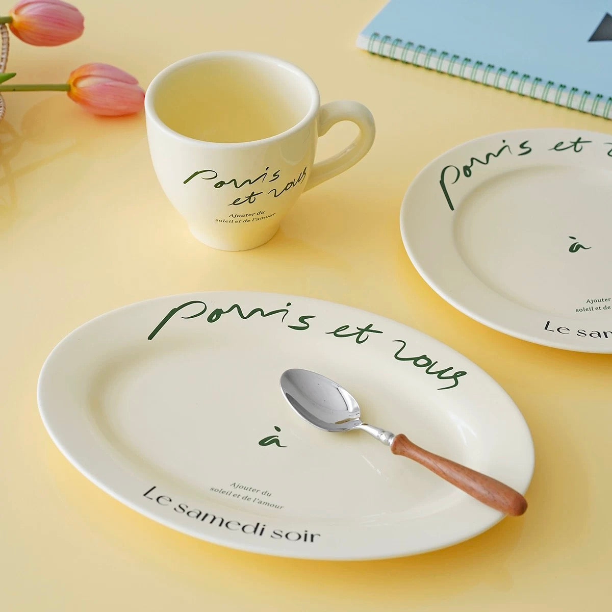 A coordinated ceramic tableware set on a pale yellow surface, including an oval plate with green script text, a matching plate partially visible at right, a white mug with French text, and a spoon with a brown handle. Tulips and a notebook appear as styled props.
