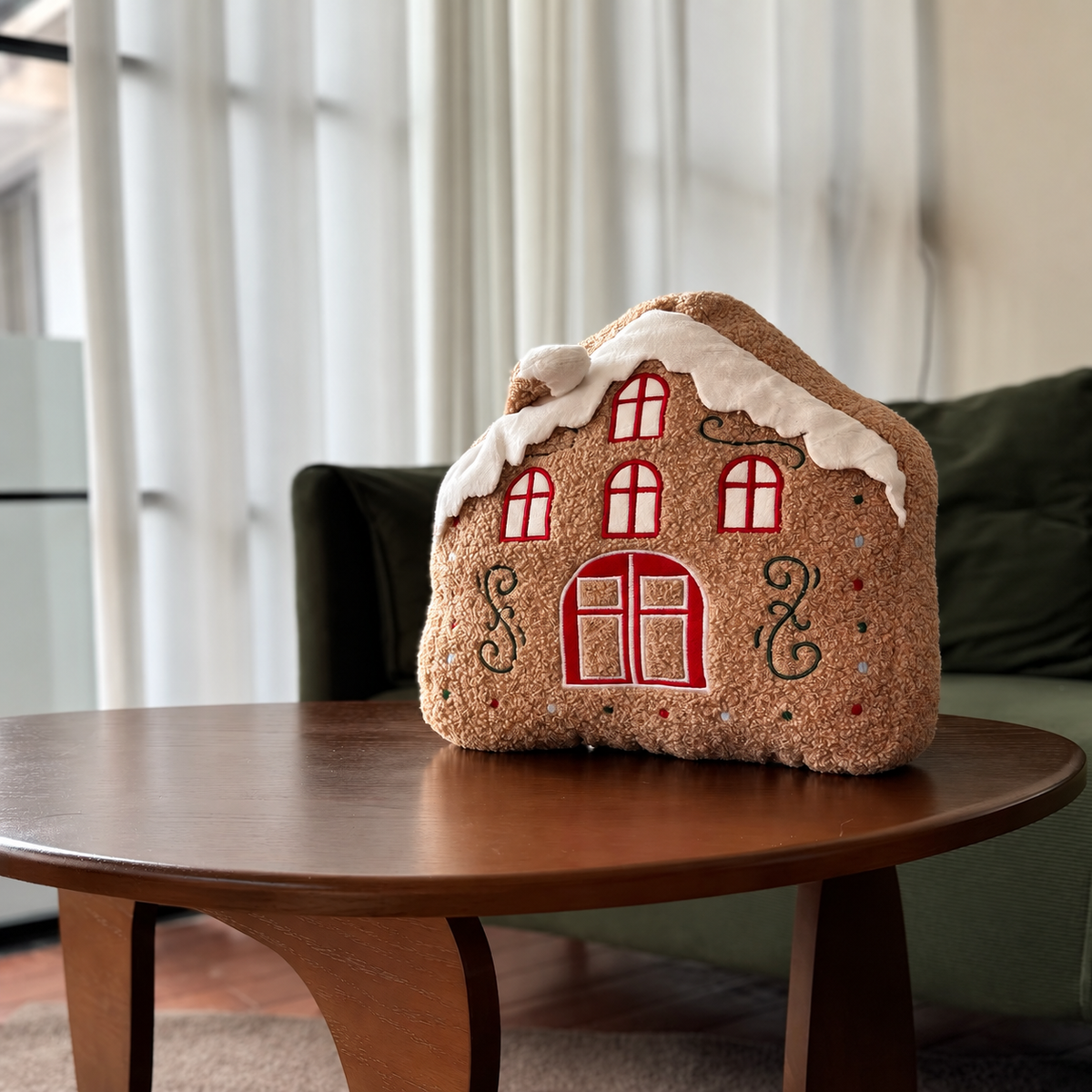 Snowy gingerbread house shaped Christmas pillow with red windows and white roof trim on a coffee table