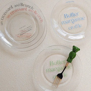 Three clear glass plates with French text are arranged on a textured cream surface. One plate has red text, one has blue text, and one has green text.