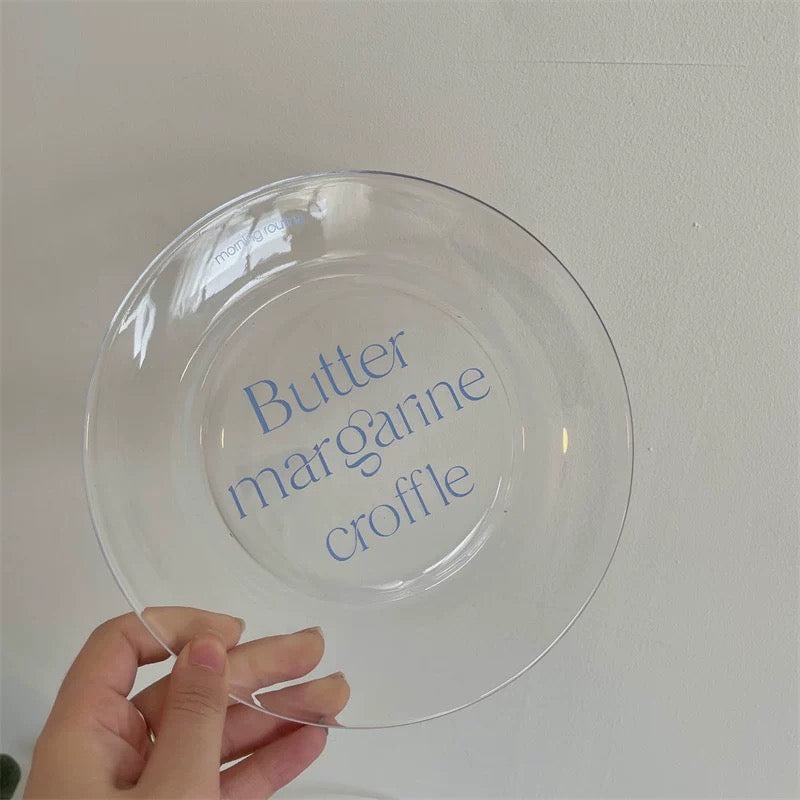 A hand holding a clear glass plate with the words 'Butter margarine croffle' printed in blue script in the center, set against a plain light gray background.