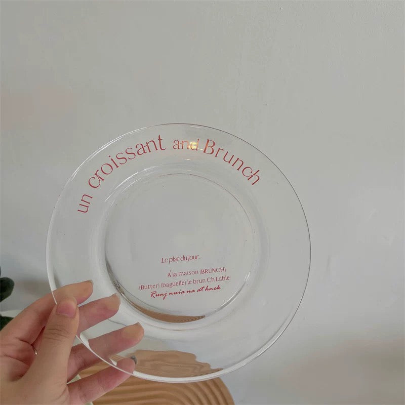 A hand holds a clear glass plate with red French text reading 'un croissant and Brunch' against a plain light gray wall.