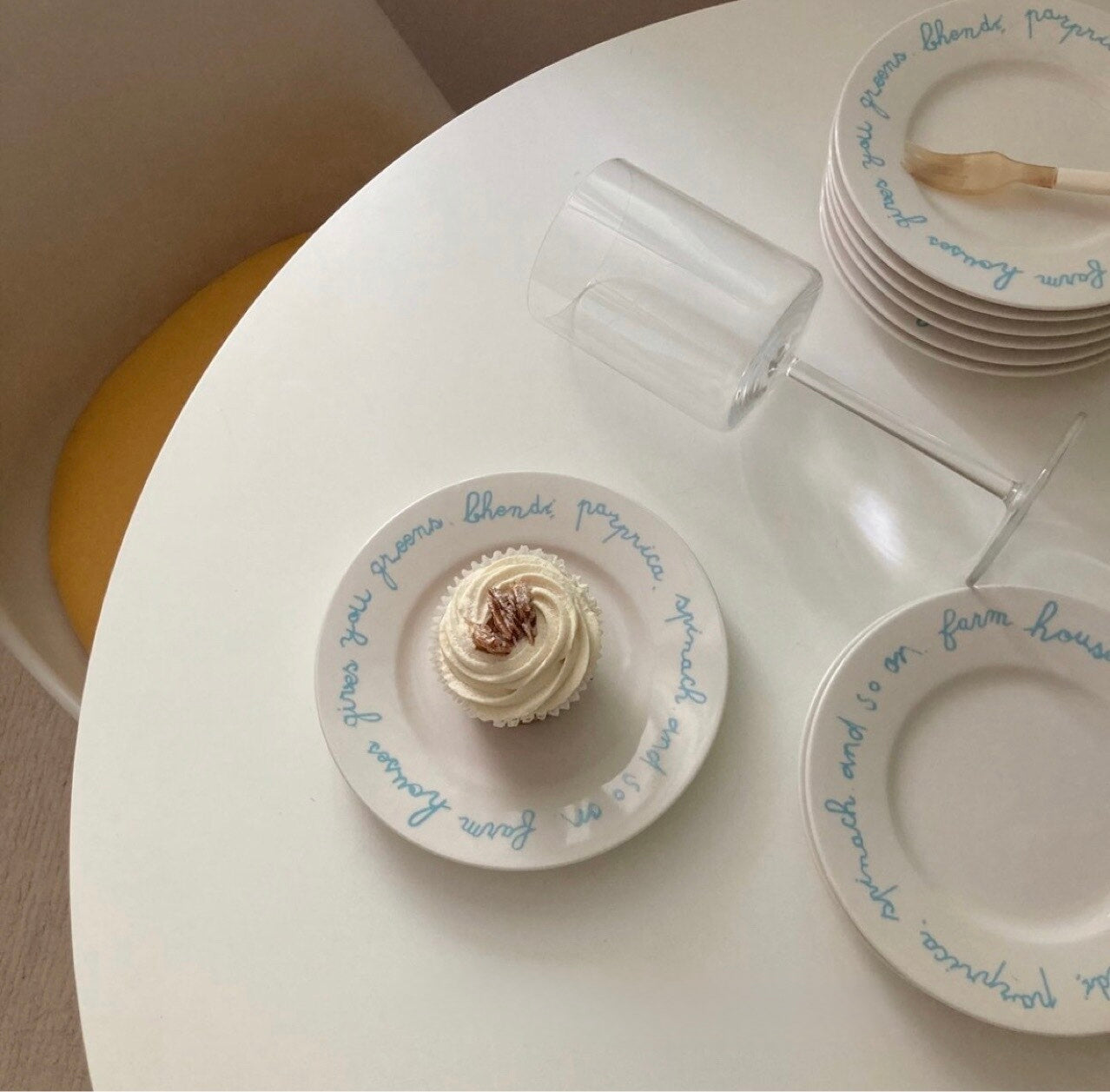 White ceramic plates with blue cursive script, shown on a round white table with a cupcake on one plate, a stack of plates, and a clear glass.