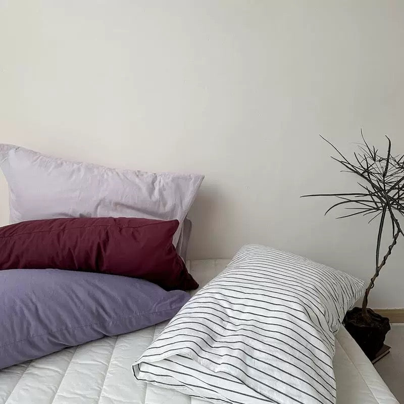 A gray pillowcase tops a stack of plum and lavender designs beside a white pillowcase with thin black stripes.