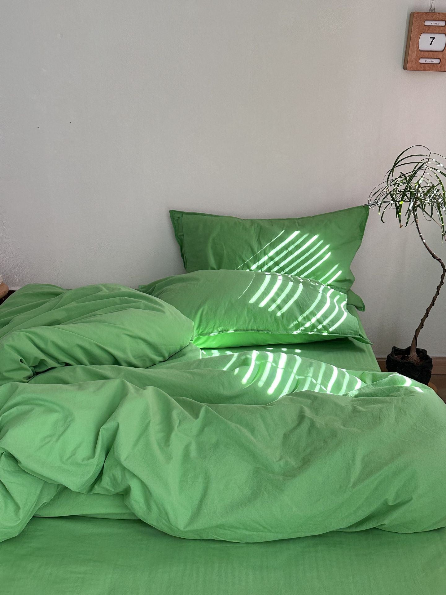 A bed made with a bright green bedding set, including a duvet cover and two matching pillowcases, styled in a minimalist bedroom with sunlight casting leaf-like shadows across the fabric.