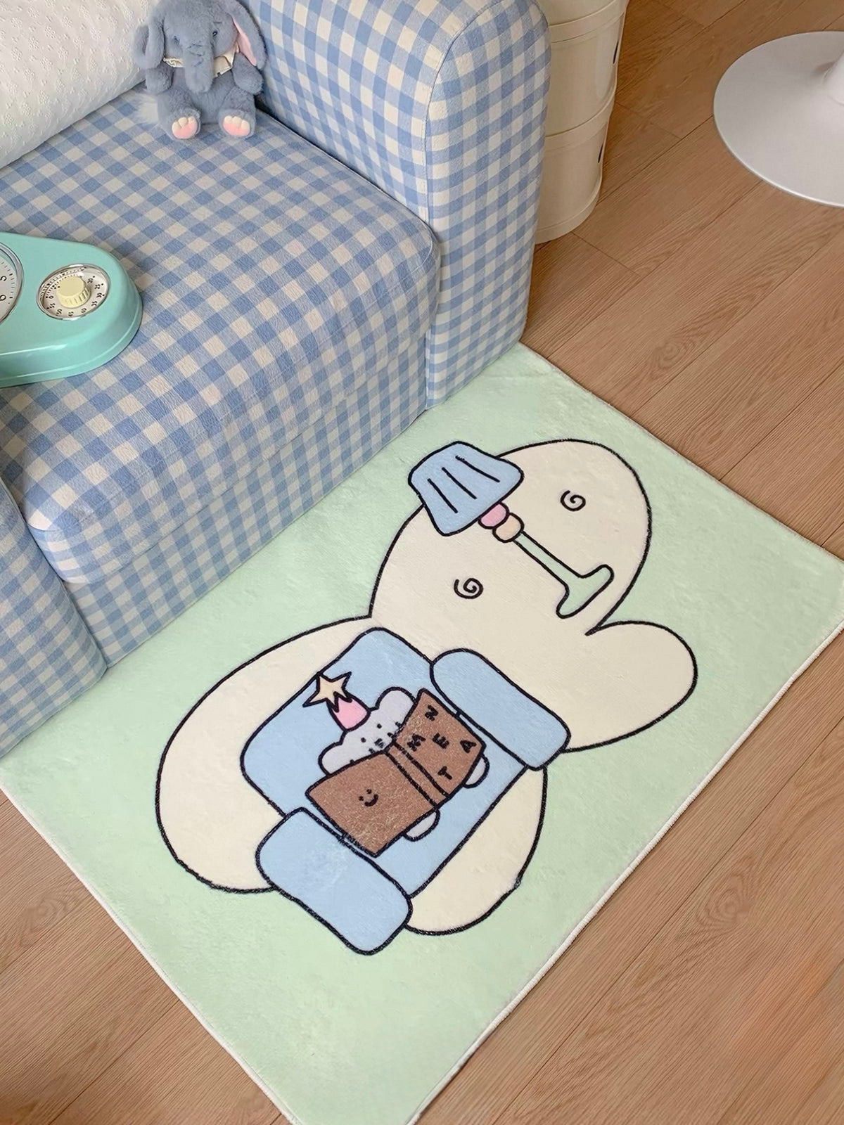 A green elephant print rug shows an elephant reading in a blue chair beside a drawn lamp, viewed at an angle.