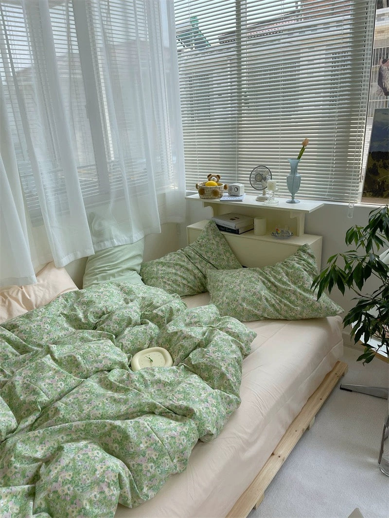 Neatly made bed with green floral bedding in a bright room with white curtains and a small table.