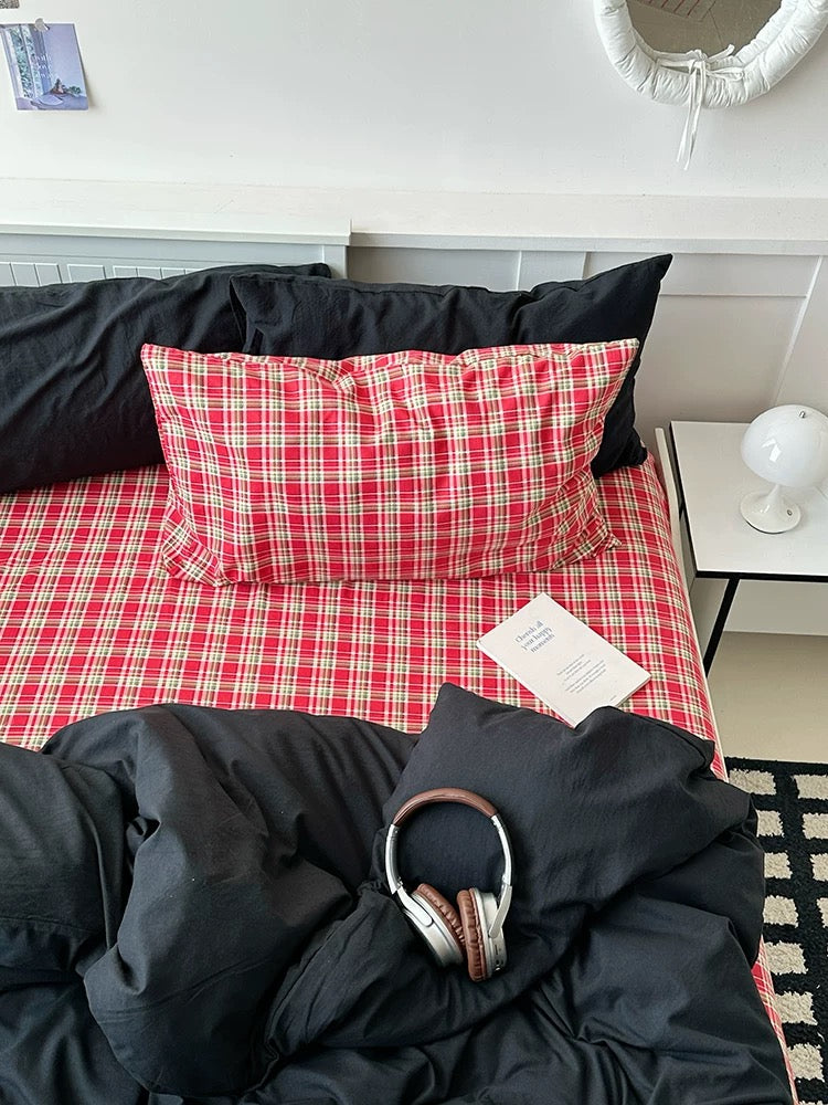 Red plaid blanket and black pillows on a bed with a person wearing headphones underneath.