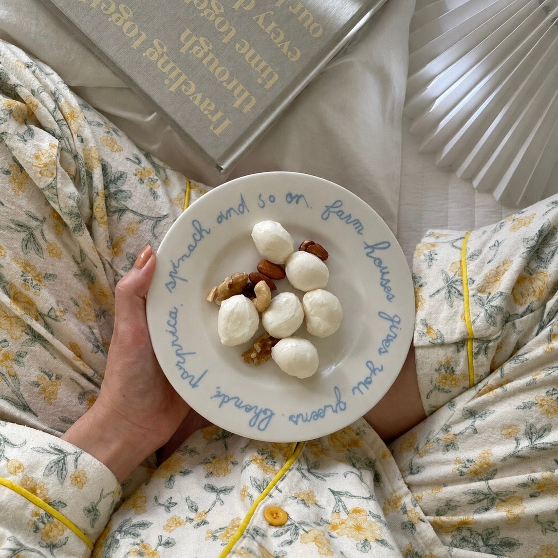 A person in floral pajamas holds a white ceramic plate with blue cursive text and small mozzarella-like cheese balls and nuts, photographed from above on a bed with a book and white fan visible.