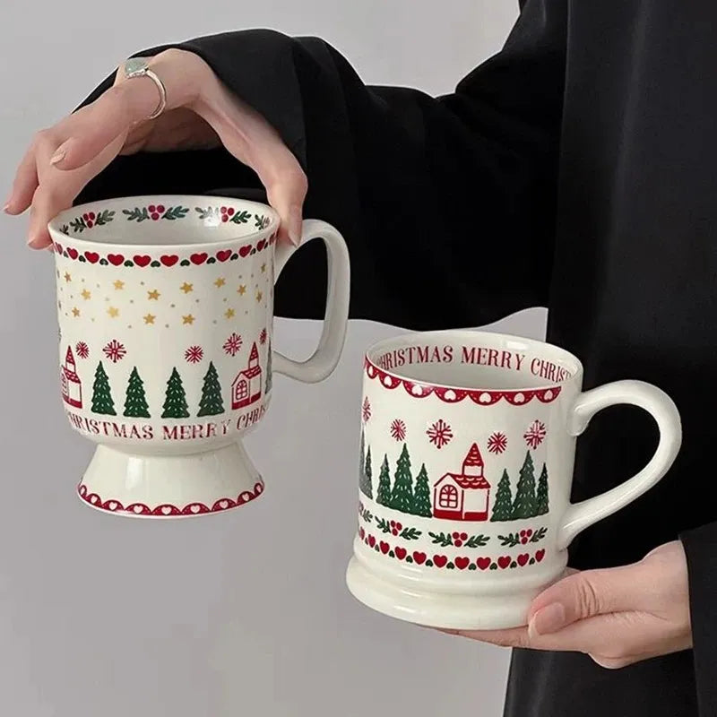 Two white ceramic Christmas-themed mugs with red and green holiday patterns, held by a person wearing a black long-sleeve top against a light gray background.