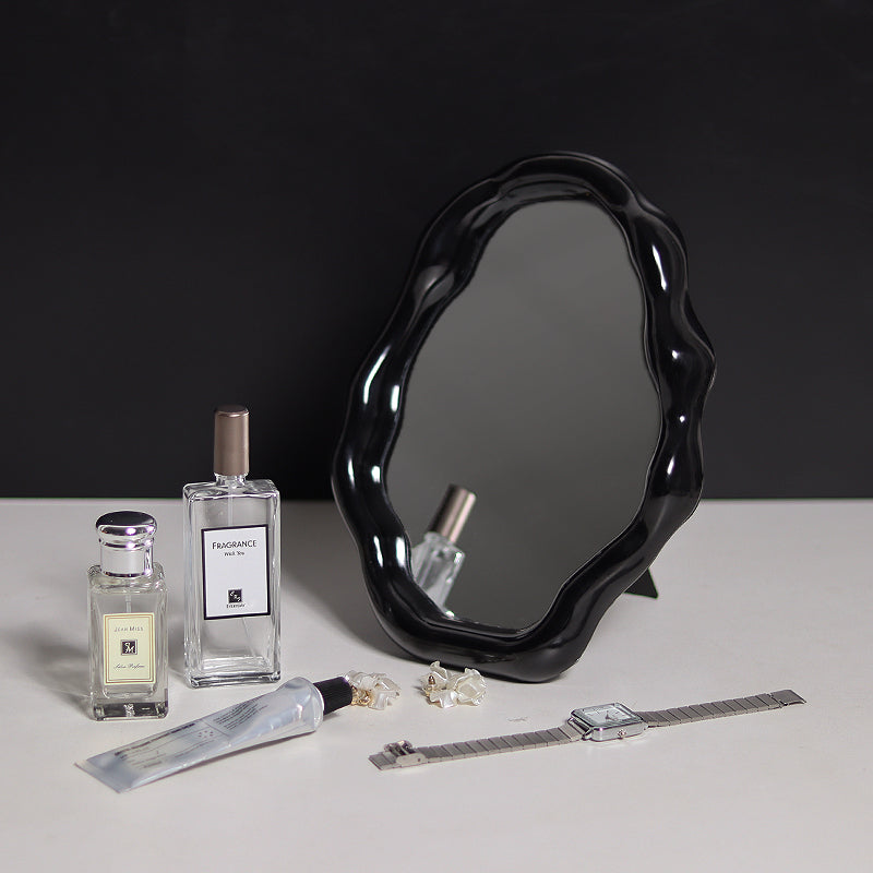 Black wavy mirror on a white surface with perfume bottles, a watch, and jewelry.